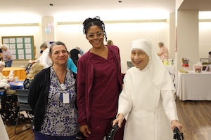 Three women smile for a photo in a brightly lit indoor community space. On the left, a woman wears a black cardigan over a blue patterned blouse and an ID badge. In the center, a woman in maroon scrubs and glasses stands tall with her hair in braids. On the right, a Catholic nun in a white habit and veil smiles while using a black walker. In the background, other people sit at tables with colorful cloths.