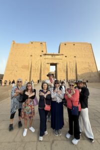 A group of six tourists standing in front of the monumental entrance pylon of the Temple of Horus at Edfu in Egypt. The women in the foreground are all posing with their arms crossed over their chests in a traditional "Pharaonic" gesture. Behind them, the massive sandstone pylon rises 36 meters high, featuring intricate ancient Egyptian reliefs of deities and pharaohs under a clear, bright sky.