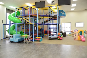 A brightly lit indoor playground features a large, multi-story climbing structure with blue and yellow framing and a tall, winding lime-green tube slide. In the foreground, there is a blue booth with a yellow table, and to the right, a smaller play area for toddlers includes a colorful plastic playhouse and a bin full of multi-colored bouncy balls. The room has high ceilings, neutral walls, and a mix of grey hard flooring and tan carpeting.