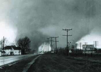 This historic 1965 black-and-white photograph, taken by Paul Huffman, captures a massive double-funnel tornado approaching the Midway Trailer Park near Dunlap, Indiana. The image shows two distinct, dark vortexes descending from a heavy cloud base over a wet road, with power lines and an "Arab Termite & Pest Control" billboard visible in the foreground.