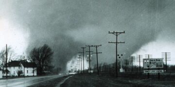 This historic 1965 black-and-white photograph, taken by Paul Huffman, captures a massive double-funnel tornado approaching the Midway Trailer Park near Dunlap, Indiana. The image shows two distinct, dark vortexes descending from a heavy cloud base over a wet road, with power lines and an "Arab Termite & Pest Control" billboard visible in the foreground.