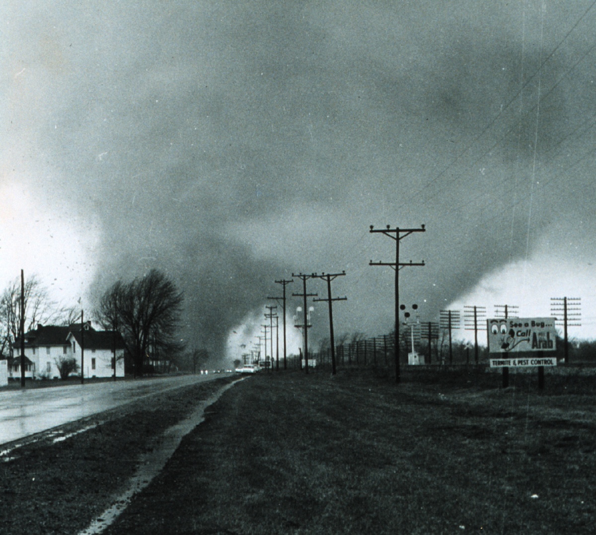 This historic 1965 black-and-white photograph, taken by Paul Huffman, captures a massive double-funnel tornado approaching the Midway Trailer Park near Dunlap, Indiana. The image shows two distinct, dark vortexes descending from a heavy cloud base over a wet road, with power lines and an "Arab Termite & Pest Control" billboard visible in the foreground.