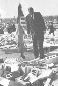 A historical black-and-white photograph from April 14, 1965, showing President Lyndon B. Johnson surveying the catastrophic damage from the Palm Sunday tornado outbreak in Indiana. He is dressed in a dark suit and stands solemnly amidst a field of splintered wood, shattered cinder blocks, and household debris. To his left, an American flag is draped over a jagged piece of upright wreckage, serving as a poignant symbol of resilience in the wake of the disaster.
