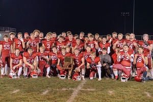 The Knox High School Redskins football team poses for a group photo on the field at night following a victory. Several players in the front row are kneeling, with one player centrally holding up the IHSAA Sectional Championship trophy. The entire team is dressed in their red and black home uniforms, celebrating together under the stadium lights.