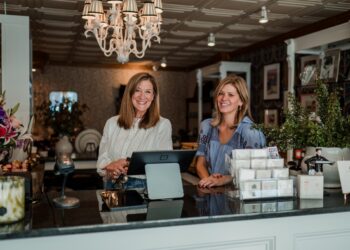 A medium shot shows two smiling women standing behind the black granite sales counter of Gifted, a boutique in Zionsville, Indiana. The woman on the left has shoulder-length brown hair and is wearing a white long-sleeved eyelet blouse; she is resting her hands on the counter near a digital point-of-sale monitor. The woman on the right, also with shoulder-length blonde hair, wears a blue patterned short-sleeved blouse. The boutique's interior features a white coffered ceiling with a large, ornate white chandelier. In the background, shelving displays various gift items, including floral arrangements, framed art, and greeting cards. To the right, a large potted plant and a stack of small gift boxes are visible on the counter.