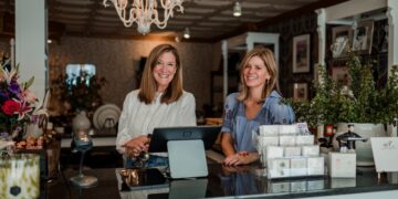 A medium shot shows two smiling women standing behind the black granite sales counter of Gifted, a boutique in Zionsville, Indiana. The woman on the left has shoulder-length brown hair and is wearing a white long-sleeved eyelet blouse; she is resting her hands on the counter near a digital point-of-sale monitor. The woman on the right, also with shoulder-length blonde hair, wears a blue patterned short-sleeved blouse. The boutique's interior features a white coffered ceiling with a large, ornate white chandelier. In the background, shelving displays various gift items, including floral arrangements, framed art, and greeting cards. To the right, a large potted plant and a stack of small gift boxes are visible on the counter.