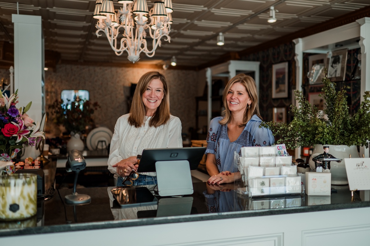 A medium shot shows two smiling women standing behind the black granite sales counter of Gifted, a boutique in Zionsville, Indiana. The woman on the left has shoulder-length brown hair and is wearing a white long-sleeved eyelet blouse; she is resting her hands on the counter near a digital point-of-sale monitor. The woman on the right, also with shoulder-length blonde hair, wears a blue patterned short-sleeved blouse. The boutique's interior features a white coffered ceiling with a large, ornate white chandelier. In the background, shelving displays various gift items, including floral arrangements, framed art, and greeting cards. To the right, a large potted plant and a stack of small gift boxes are visible on the counter.