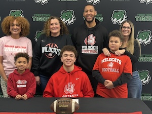 A group of seven people—three adults and four children—pose for a celebratory photo behind a table with a football and a signing paper. Everyone is wearing "Wildcats Football" or "Wildcats" apparel in red, pink, and black. They are standing in front of a dark backdrop featuring the Yorktown Tigers green tiger head logo.