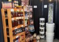 An indoor market display featuring locally sourced products. A wooden shelf is stocked with jars of Danny's Maple syrup, honey, and colorful bottled drinks. Next to it, a wall display showcases various flavors of beef jerky and meat snacks. Large white buckets and ceramic jugs are stacked on the floor against a black backdrop decorated with warm string lights.