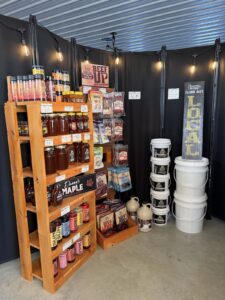 An indoor market display featuring locally sourced products. A wooden shelf is stocked with jars of Danny's Maple syrup, honey, and colorful bottled drinks. Next to it, a wall display showcases various flavors of beef jerky and meat snacks. Large white buckets and ceramic jugs are stacked on the floor against a black backdrop decorated with warm string lights.