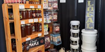 An indoor market display featuring locally sourced products. A wooden shelf is stocked with jars of Danny's Maple syrup, honey, and colorful bottled drinks. Next to it, a wall display showcases various flavors of beef jerky and meat snacks. Large white buckets and ceramic jugs are stacked on the floor against a black backdrop decorated with warm string lights.