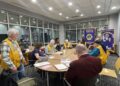 A group of about ten people are gathered around a large, light-wood oval table in a modern meeting room with grey carpet and large windows. Most of the participants are wearing bright yellow vests with blue trim and patches, identifying them as members of the Plainfield Lions Club. In the background, two purple and gold banners are prominently displayed, one reading "Plainfield Lions Club, Indiana" and the other showcasing various club awards. Papers and folders are scattered across the table as the group engages in a discussion.