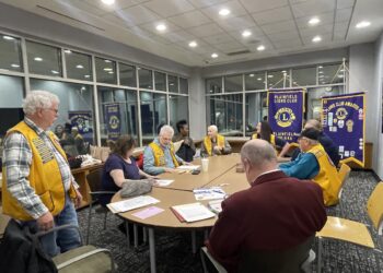 A group of about ten people are gathered around a large, light-wood oval table in a modern meeting room with grey carpet and large windows. Most of the participants are wearing bright yellow vests with blue trim and patches, identifying them as members of the Plainfield Lions Club. In the background, two purple and gold banners are prominently displayed, one reading "Plainfield Lions Club, Indiana" and the other showcasing various club awards. Papers and folders are scattered across the table as the group engages in a discussion.