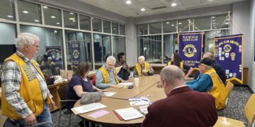 A group of about ten people are gathered around a large, light-wood oval table in a modern meeting room with grey carpet and large windows. Most of the participants are wearing bright yellow vests with blue trim and patches, identifying them as members of the Plainfield Lions Club. In the background, two purple and gold banners are prominently displayed, one reading "Plainfield Lions Club, Indiana" and the other showcasing various club awards. Papers and folders are scattered across the table as the group engages in a discussion.