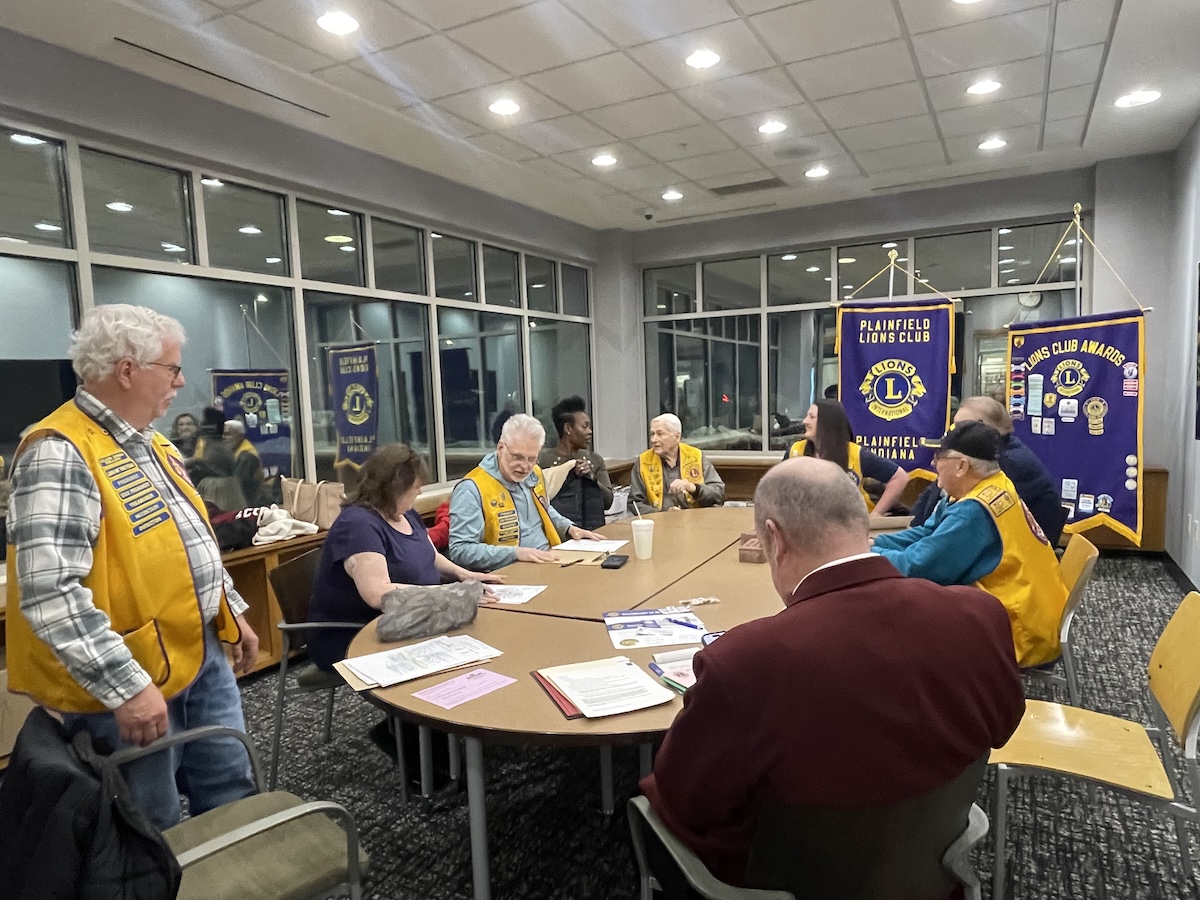 A group of about ten people are gathered around a large, light-wood oval table in a modern meeting room with grey carpet and large windows. Most of the participants are wearing bright yellow vests with blue trim and patches, identifying them as members of the Plainfield Lions Club. In the background, two purple and gold banners are prominently displayed, one reading "Plainfield Lions Club, Indiana" and the other showcasing various club awards. Papers and folders are scattered across the table as the group engages in a discussion.