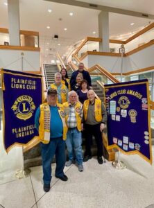 Members of the Plainfield Lions Club pose for a group photo on a modern, multi-tiered staircase inside the MADE@Plainfield facility. The group, consisting of seven adults, is positioned on the stairs, with three men in the front row wearing signature yellow Lions Club vests. They are flanked by two large purple and gold banners: one on the left identifies the "Plainfield Lions Club, Plainfield, Indiana," and the one on the right displays various "Lions Club Awards" and patches. The background features the building's bright, open lobby with white pillars, glass railings, and light wood accents.