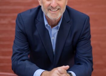 A professional studio headshot of John Stehr, the Mayor of Zionsville, Indiana. He is a man with short, silver hair and a warm, approachable smile. He is wearing a dark navy blue blazer over a light blue button-down shirt. He is leaning forward with his forearms resting on a horizontal red railing, and his hands are loosely clasped in front of him. The background is a softly blurred red brick wall, which provides a classic and urban texture to the portrait.