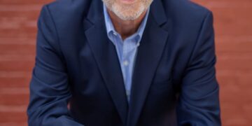 A professional studio headshot of John Stehr, the Mayor of Zionsville, Indiana. He is a man with short, silver hair and a warm, approachable smile. He is wearing a dark navy blue blazer over a light blue button-down shirt. He is leaning forward with his forearms resting on a horizontal red railing, and his hands are loosely clasped in front of him. The background is a softly blurred red brick wall, which provides a classic and urban texture to the portrait.