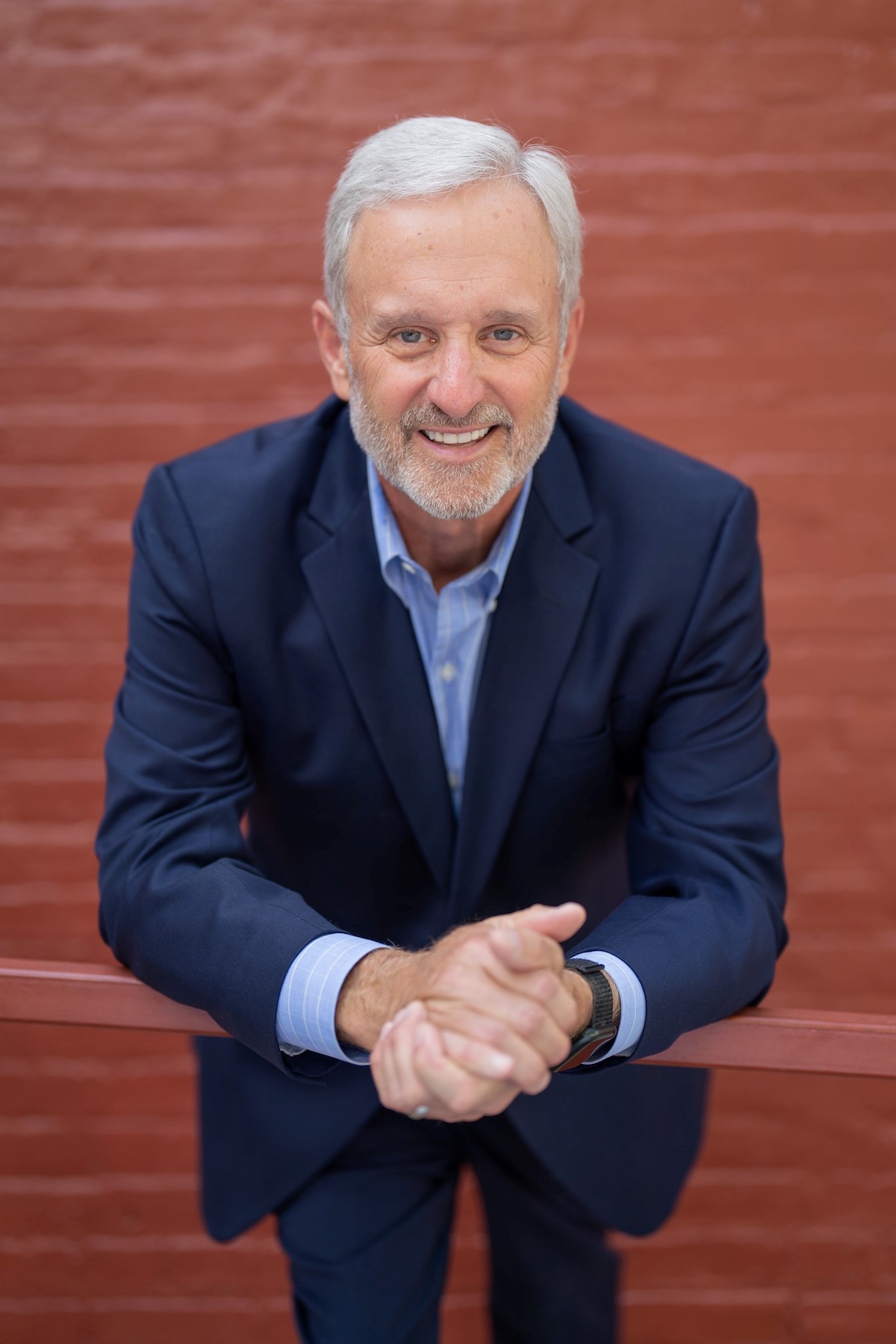 A professional studio headshot of John Stehr, the Mayor of Zionsville, Indiana. He is a man with short, silver hair and a warm, approachable smile. He is wearing a dark navy blue blazer over a light blue button-down shirt. He is leaning forward with his forearms resting on a horizontal red railing, and his hands are loosely clasped in front of him. The background is a softly blurred red brick wall, which provides a classic and urban texture to the portrait.