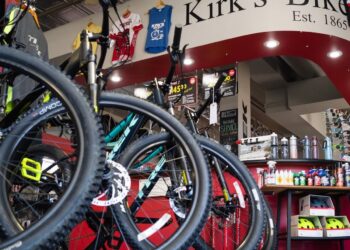 A low-angle view of the interior of Kirk's Bike Shop, featuring rows of Trek mountain bikes with prominent black tires and disc brakes in the foreground. A large, curved white header sign reading "Kirk's Bike Est. 1865" spans the ceiling. In the background, a red-walled service and accessory area is stocked with water bottles and cycling supplies, while colorful jerseys hang from the ceiling.