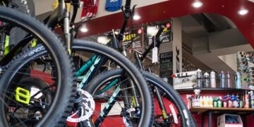 A low-angle view of the interior of Kirk's Bike Shop, featuring rows of Trek mountain bikes with prominent black tires and disc brakes in the foreground. A large, curved white header sign reading "Kirk's Bike Est. 1865" spans the ceiling. In the background, a red-walled service and accessory area is stocked with water bottles and cycling supplies, while colorful jerseys hang from the ceiling.