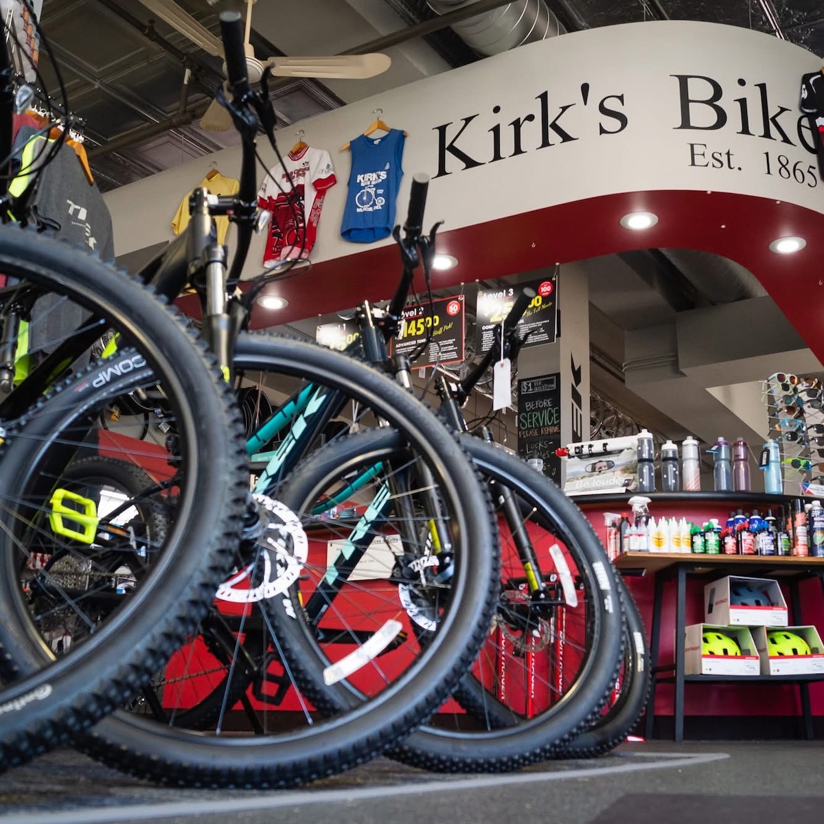 A low-angle view of the interior of Kirk's Bike Shop, featuring rows of Trek mountain bikes with prominent black tires and disc brakes in the foreground. A large, curved white header sign reading "Kirk's Bike Est. 1865" spans the ceiling. In the background, a red-walled service and accessory area is stocked with water bottles and cycling supplies, while colorful jerseys hang from the ceiling.