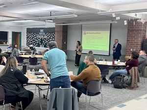 A medium shot captures a community or school board presentation in progress at Westfield High School. A female presenter stands near a large projector screen displaying a bright yellow slide with the title "Destination Westfield" and several bullet points about district goals. To the right, Westfield Washington Schools Superintendent Dr. John Atha, dressed in a navy suit, watches the presentation. The room, which has a mix of white and red brick walls, is filled with several circular wooden tables where community members and staff are seated, some looking at the screen and others in conversation.

