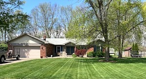 A wide, horizontal shot of a well-maintained ranch-style home in a suburban neighborhood. The house features a mix of light tan siding and red brick accents, with a large two-car garage on the left and a prominent gabled entryway. A large, mature deciduous tree with fresh green spring leaves stands in the front yard to the right. The focal point of the image is the expansive, lush green lawn, which is neatly mowed into a distinct vertical striped pattern. A light-colored concrete driveway leads to the garage, where a portion of a vehicle and trailer are visible. Other mature trees and a small wooden shed are seen in the soft-focus background under a clear blue sky.