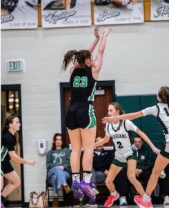 A high-action shot of a high school basketball game. A player wearing a black jersey with green accents and the number 23 jumps high in the air to take a shot. An opposing player in a white "Arabians" jersey with the number 2 tries to defend. In the background, spectators sit in the stands below championship banners for the class of 2024.