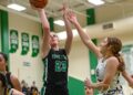 A high-action shot of a high school basketball game. A player from Yorktown, wearing a black jersey with the number 23, jumps to take a shot, with the ball poised high above her head. An opposing player in a white jersey reaches up with one hand in an attempt to block the shot. In the background, other players, spectators, and green championship banners are visible in the brightly lit gymnasium.