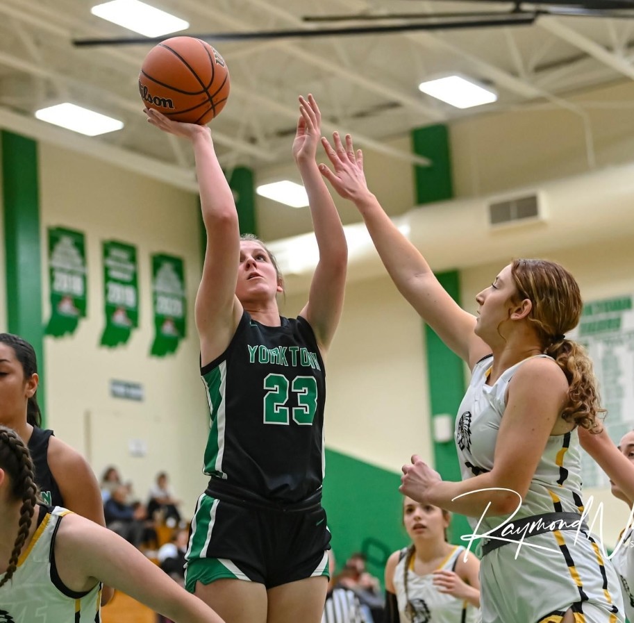 A high-action shot of a high school basketball game. A player from Yorktown, wearing a black jersey with the number 23, jumps to take a shot, with the ball poised high above her head. An opposing player in a white jersey reaches up with one hand in an attempt to block the shot. In the background, other players, spectators, and green championship banners are visible in the brightly lit gymnasium.