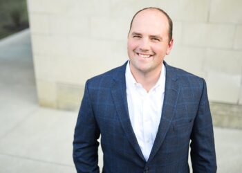 A professional headshot of Scott Fadness, the Mayor of Fishers, Indiana, smiling warmly at the camera. He is a man with short brown hair and is wearing a dark blue, faint windowpane-check blazer over a crisp white button-down shirt. He is outdoors, positioned in front of a light-colored, modern stone building with a concrete walkway visible in the soft-focus background. The lighting is bright and even, highlighting a friendly and approachable expression.