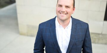 A professional headshot of Scott Fadness, the Mayor of Fishers, Indiana, smiling warmly at the camera. He is a man with short brown hair and is wearing a dark blue, faint windowpane-check blazer over a crisp white button-down shirt. He is outdoors, positioned in front of a light-colored, modern stone building with a concrete walkway visible in the soft-focus background. The lighting is bright and even, highlighting a friendly and approachable expression.