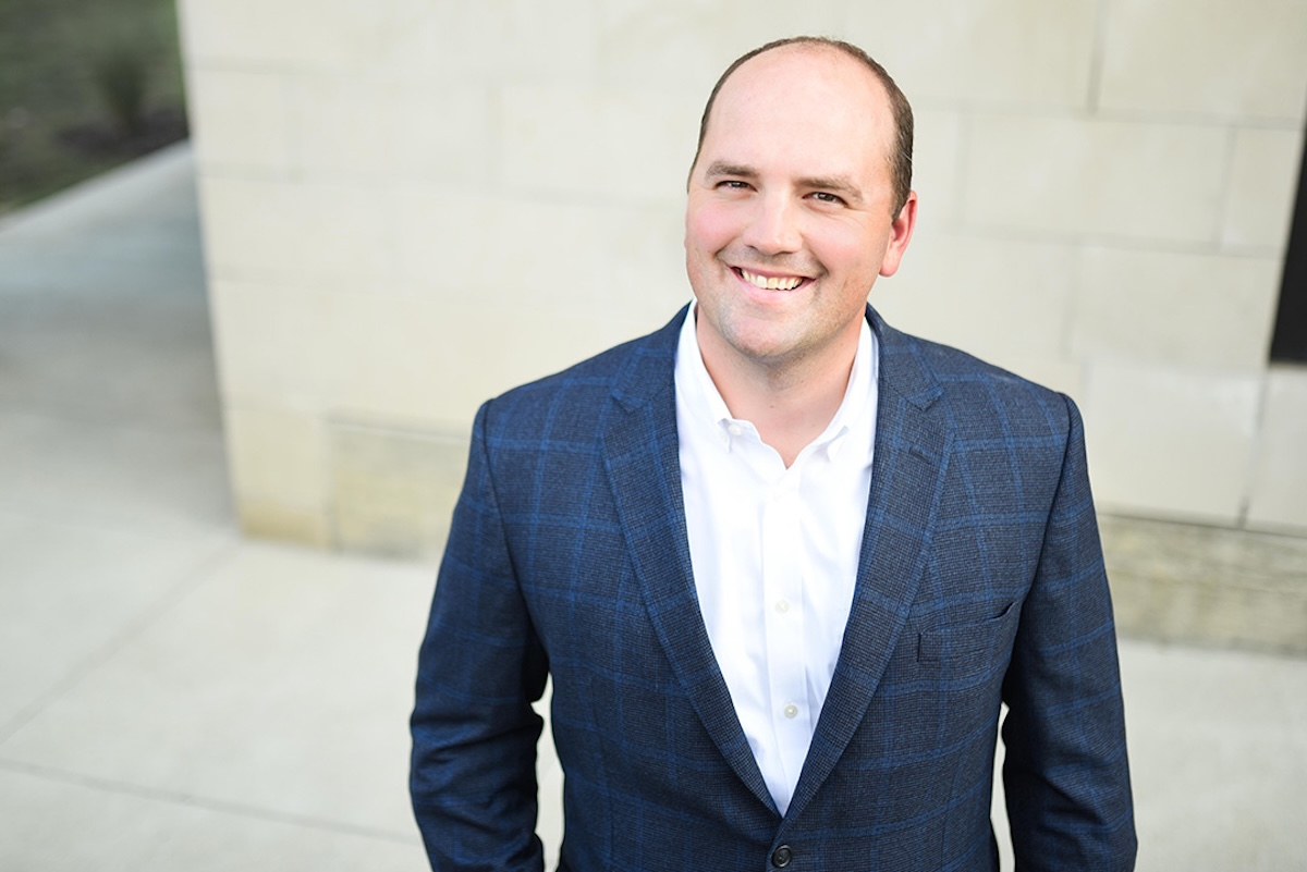 A professional headshot of Scott Fadness, the Mayor of Fishers, Indiana, smiling warmly at the camera. He is a man with short brown hair and is wearing a dark blue, faint windowpane-check blazer over a crisp white button-down shirt. He is outdoors, positioned in front of a light-colored, modern stone building with a concrete walkway visible in the soft-focus background. The lighting is bright and even, highlighting a friendly and approachable expression.