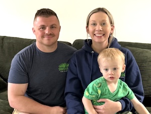 A warm family portrait of a man, a woman, and a young child sitting together on a dark gray corduroy sofa. The man on the left wears a charcoal gray t-shirt with a green "Southside Turf Solutions" logo on the chest. The woman in the center wears a navy blue hoodie with the same logo and is smiling broadly. She has her arm around the young boy sitting on her lap, who is looking slightly away from the camera and wearing a bright green t-shirt. The background is a plain white wall, creating a clean and personal feel.