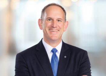 A professional headshot of a smiling man with short, thinning brown hair, posed with his arms crossed. He is wearing a dark navy blue suit jacket, a crisp white button-down shirt, and a solid bright blue silk tie. He also has a small, diamond-shaped silver lapel pin. The background is a brightly lit, modern office space with a large wall of glowing, out-of-focus circular lights, creating a soft bokeh effect.