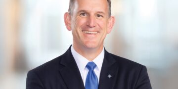 A professional headshot of a smiling man with short, thinning brown hair, posed with his arms crossed. He is wearing a dark navy blue suit jacket, a crisp white button-down shirt, and a solid bright blue silk tie. He also has a small, diamond-shaped silver lapel pin. The background is a brightly lit, modern office space with a large wall of glowing, out-of-focus circular lights, creating a soft bokeh effect.