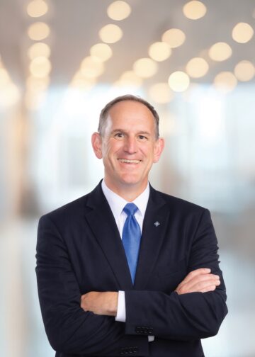 A professional headshot of a smiling man with short, thinning brown hair, posed with his arms crossed. He is wearing a dark navy blue suit jacket, a crisp white button-down shirt, and a solid bright blue silk tie. He also has a small, diamond-shaped silver lapel pin. The background is a brightly lit, modern office space with a large wall of glowing, out-of-focus circular lights, creating a soft bokeh effect.