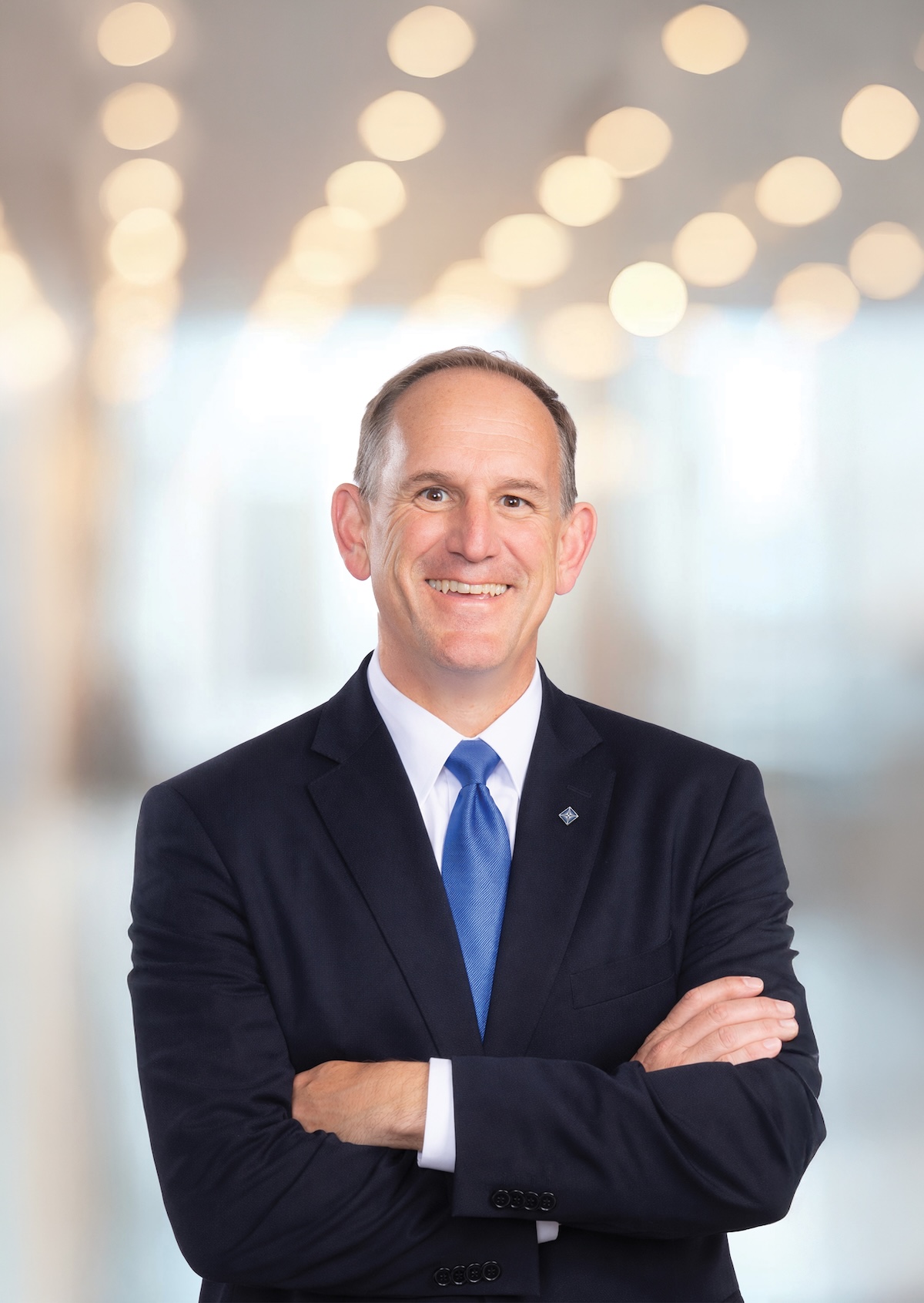 A professional headshot of a smiling man with short, thinning brown hair, posed with his arms crossed. He is wearing a dark navy blue suit jacket, a crisp white button-down shirt, and a solid bright blue silk tie. He also has a small, diamond-shaped silver lapel pin. The background is a brightly lit, modern office space with a large wall of glowing, out-of-focus circular lights, creating a soft bokeh effect.