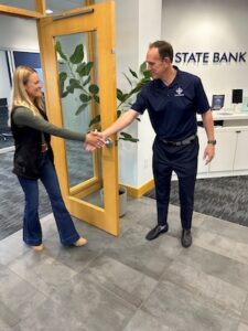 Inside a modern bank lobby, a man and a woman are smiling and shaking hands in front of an open light-wood door with glass panes. The man, on the right, wears a navy blue polo shirt with a white circular logo, navy trousers, and black dress shoes. The woman, on the left, wears an olive green long-sleeved shirt, a black vest, and dark blue flared jeans. In the background, a white wall features the partial text "STATE BANK" in bold, dark blue capital letters.