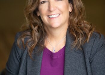 A professional studio headshot of a woman with shoulder-length, wavy brown hair and brown eyes, smiling warmly at the camera. She is wearing a purple top under a textured dark gray blazer, accented by a delicate silver necklace with a small pendant. The portrait is taken with a shallow depth of field, making the dark, neutral-toned background softly blurred and focusing entirely on her friendly and professional expression.