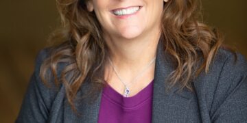 A professional studio headshot of a woman with shoulder-length, wavy brown hair and brown eyes, smiling warmly at the camera. She is wearing a purple top under a textured dark gray blazer, accented by a delicate silver necklace with a small pendant. The portrait is taken with a shallow depth of field, making the dark, neutral-toned background softly blurred and focusing entirely on her friendly and professional expression.