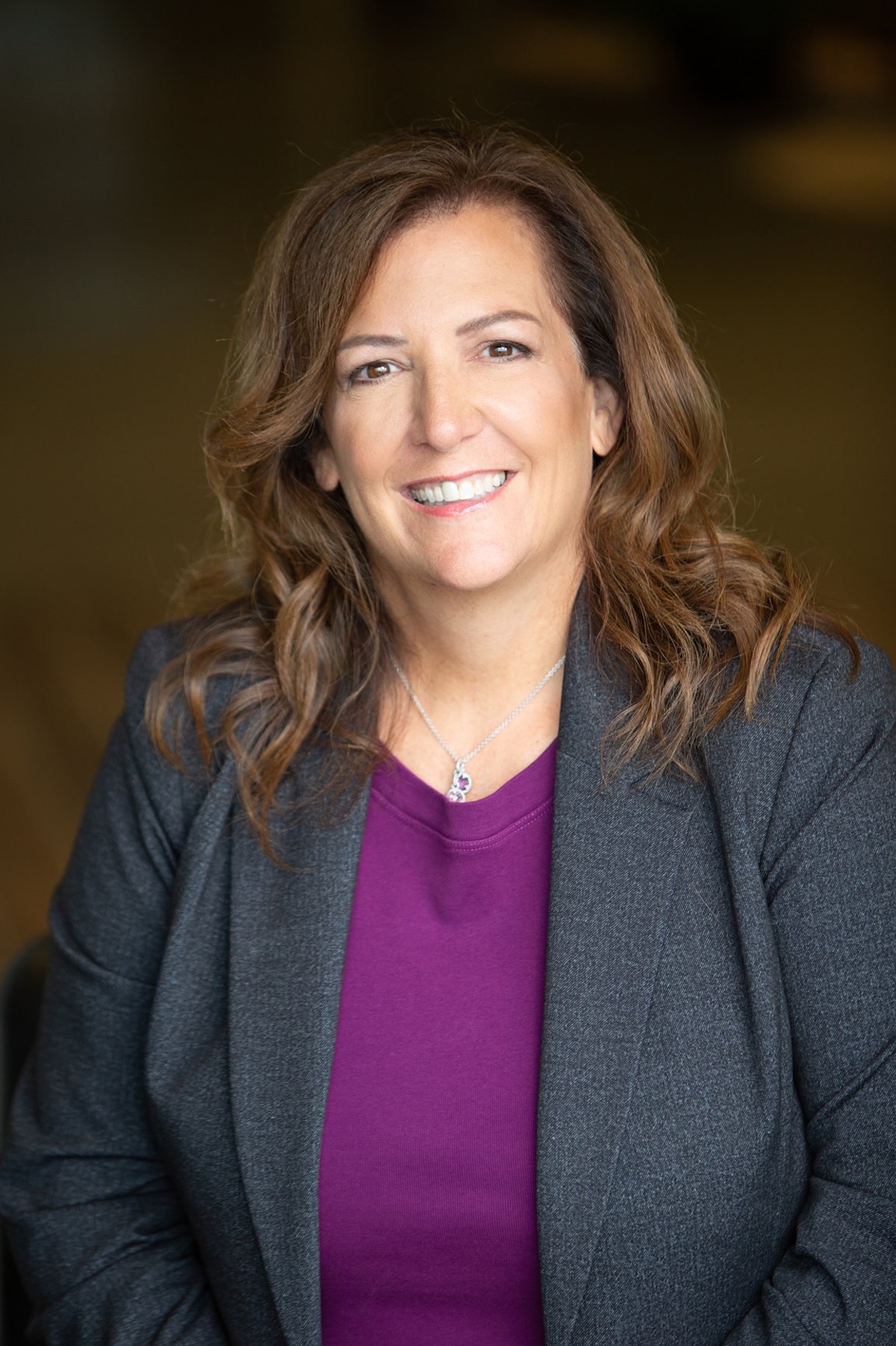 A professional studio headshot of a woman with shoulder-length, wavy brown hair and brown eyes, smiling warmly at the camera. She is wearing a purple top under a textured dark gray blazer, accented by a delicate silver necklace with a small pendant. The portrait is taken with a shallow depth of field, making the dark, neutral-toned background softly blurred and focusing entirely on her friendly and professional expression.
