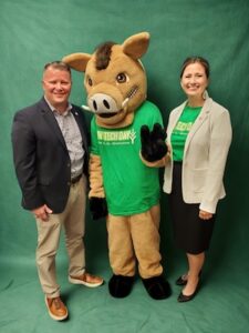 A medium shot of three people posing together in front of a solid green backdrop for Ivy Tech Day 2026. On the left, a man in a navy blue blazer over a checkered button-down shirt and tan chinos smiles at the camera. In the center is Boris the Wild Boar, the mascot for Ivy Tech Hamilton County, wearing a bright green t-shirt with the "IVY TECH DAY" logo. Boris has one black-gloved hand raised in a wave. On the right, Dr. Rachel Kartz, Chancellor of the Hamilton County campus, smiles warmly; she is wearing a light beige blazer over a matching green "IVY TECH DAY" t-shirt and a black skirt.