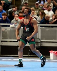 An action shot captures the intense celebration of Michael White (#190) from Lawrence North High School at the 2026 IHSAA Boys Wrestling State Finals. He is standing on a light blue wrestling mat at Gainbridge Fieldhouse, his body tensed and muscles flexed in a triumphant pose. He is shouting with joy, his mouth open and head tilted back. He wears a black, red, and green "LN" wrestling singlet, black wrestling shoes, and headgear pushed up on his forehead. In the background, a large, blurred crowd of spectators watches from the stands behind a metal railing.