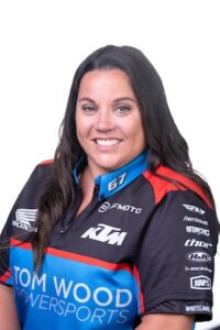 A professional studio headshot of Mindy Layton, General Manager of Tom Wood Powersports South. She has long, wavy dark hair and is smiling warmly at the camera against a plain white background. She is wearing a custom black, blue, and red zippered team jersey that features prominent logos for Tom Wood Powersports, KTM, and CFMOTO on the front. The left sleeve displays several other industry brand logos, including Fly Racing, Fasthouse, Stacyc, Thor, HJC, Dunlop, and 100%, with "WHITELAND, IN" printed at the bottom of the sleeve.