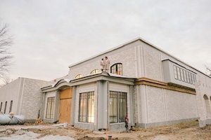 A wide, low-angle shot of the Westwood Vowery & Estate wedding venue under construction in Westfield, Indiana. The large, modern limestone-textured building features grand arched windows and a prominent entryway with a temporary wooden door. Two people, Shelby and Seth Dempsey, are standing on a second-story rooftop terrace overlooking the property. The surrounding ground is covered in dirt and construction debris, with a large white propane tank visible on the left. The sky is a soft, overcast gray, emphasizing the scale and clean lines of the 17,000-square-foot facility.