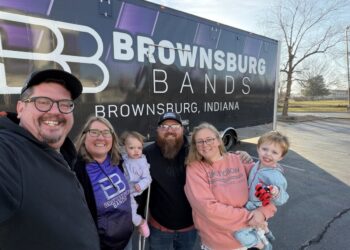 A group of five adults and two young children pose for a smiling selfie in a sunny parking lot. They are standing in front of a large, black semi-truck trailer with purple and white branding that reads "BROWNSBURG BANDS" and "BROWNSBURG, INDIANA." One woman wears a purple hoodie with the "BB" logo, and the group appears to be a family or group of dedicated supporters.