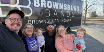 A group of five adults and two young children pose for a smiling selfie in a sunny parking lot. They are standing in front of a large, black semi-truck trailer with purple and white branding that reads "BROWNSBURG BANDS" and "BROWNSBURG, INDIANA." One woman wears a purple hoodie with the "BB" logo, and the group appears to be a family or group of dedicated supporters.