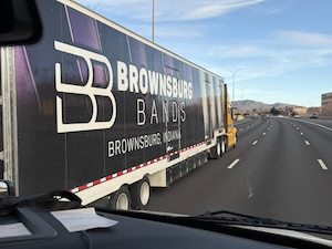 A point-of-view shot from the dashboard of a vehicle driving on a multi-lane highway, showing a large semi-truck in the next lane. The truck's black trailer features prominent white and purple branding for "BROWNSBURG BANDS" from "BROWNSBURG, INDIANA," along with a stylized "BB" logo. Mountains are visible in the distant background under a bright, clear blue sky.